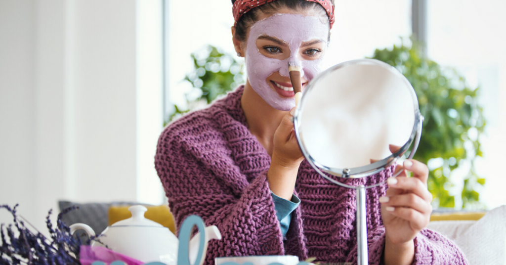 Mujer joven que aplica la máscara facial. Está sentada en su cama mirando al espejo y cuidando su piel durante la mañana del fin de semana.