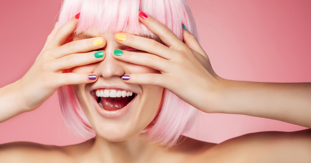 una chica se tapa los ojos y se rie lleva las uñas pintadas cada una de un color rosa amarillo verde y azul el pelo es rosa y corto con flequillo