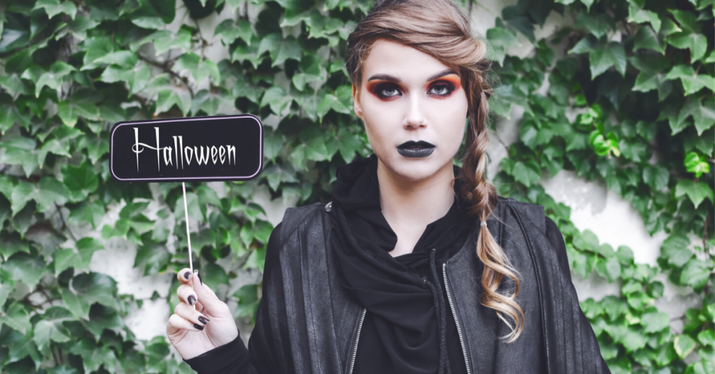 chica joven con los labios pintados de negro, los ojos con sombras y la cara blanca lleva una trenza al lado y sostiene vestida de negro un cartel que dice halloween