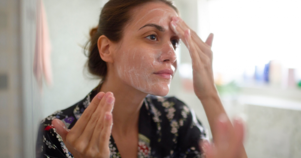mujer joven se limpia la cara con las manos y una espuma blanca frente al espejo de un cuarto de baño