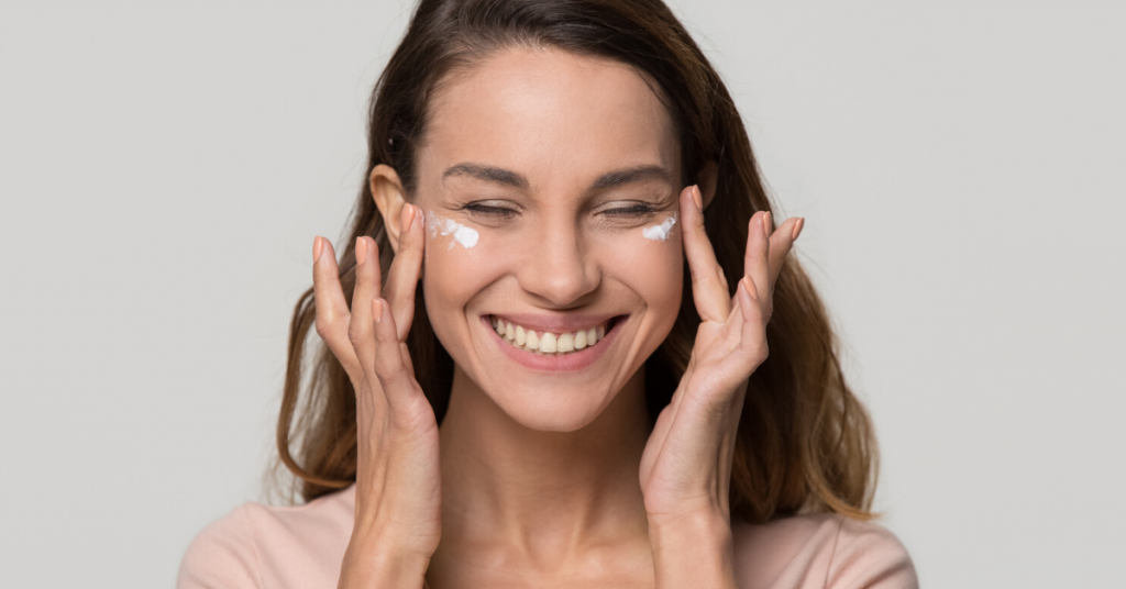 Chica bastante joven sonríe feliz mientras se aplica crema cosmética loción hidratante en la cara para el cuidado de la piel limpia suave y saludable tratamiento de belleza natural aislado sobre fondo gris blanco de estudio