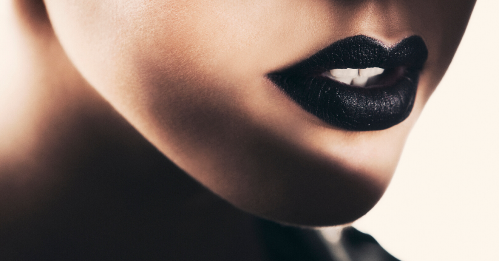 foto detalle de los labios de una mujer joven pintados con carmin negro oscuro. Una sombra cubre parte de la barbilla y el cuello.