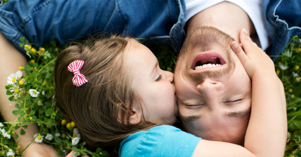 padre joven tiumbado en la hierba es besado por su hija pequeña en la cara mientras él se rie